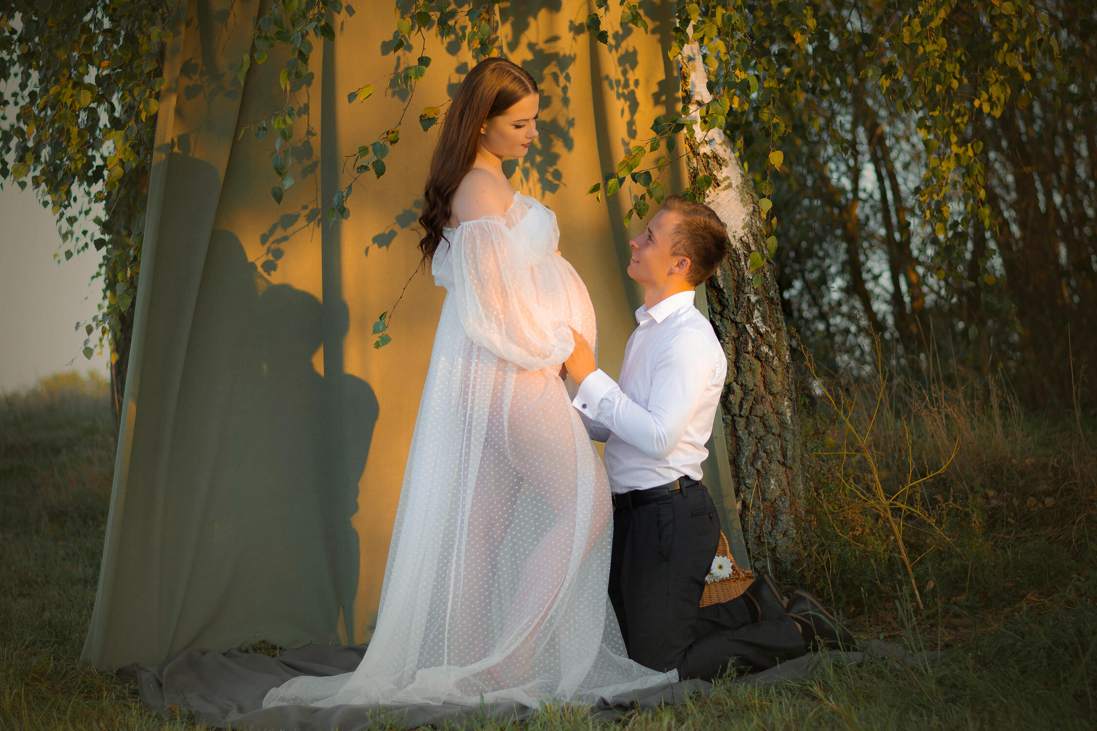 Nėštukės fotosesija gamtoje, vyras klūpi ant kelių ir laiko nėščiosios pilvuką