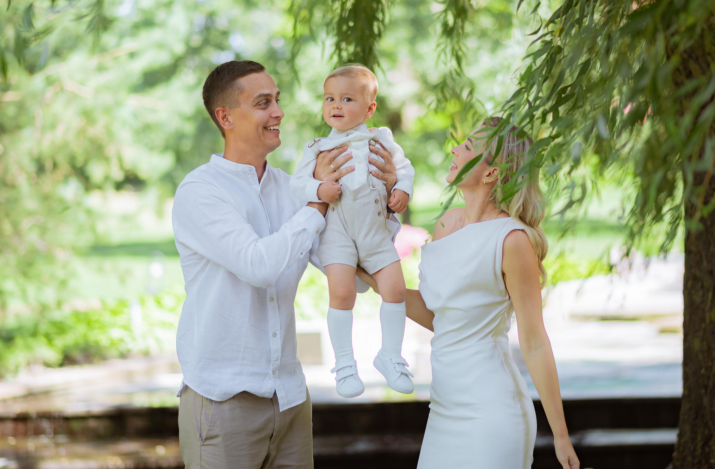 Šeimos fotografė įamžina tėvus, žaidžiančius su vaiku parke. Natūrali šeimos fotosesija lauke, skirta išsaugoti vaikystės prisiminimus, šeimos bendrystę ir spontaniškas emocijas