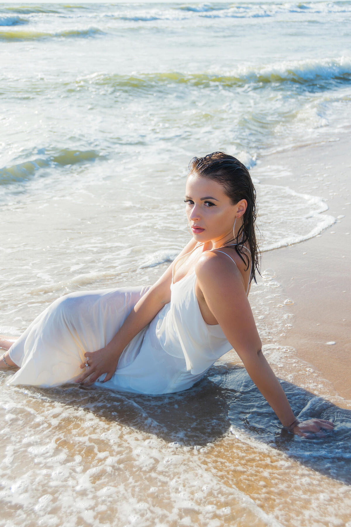 Asmeninė fotosesija atlikta svečiuojantis pajūryje Jauna moteris balta suknele pajūryje sėdi bangose – vasariška fotosesija prie jūros. Profesionali fotosesija gamtoje.