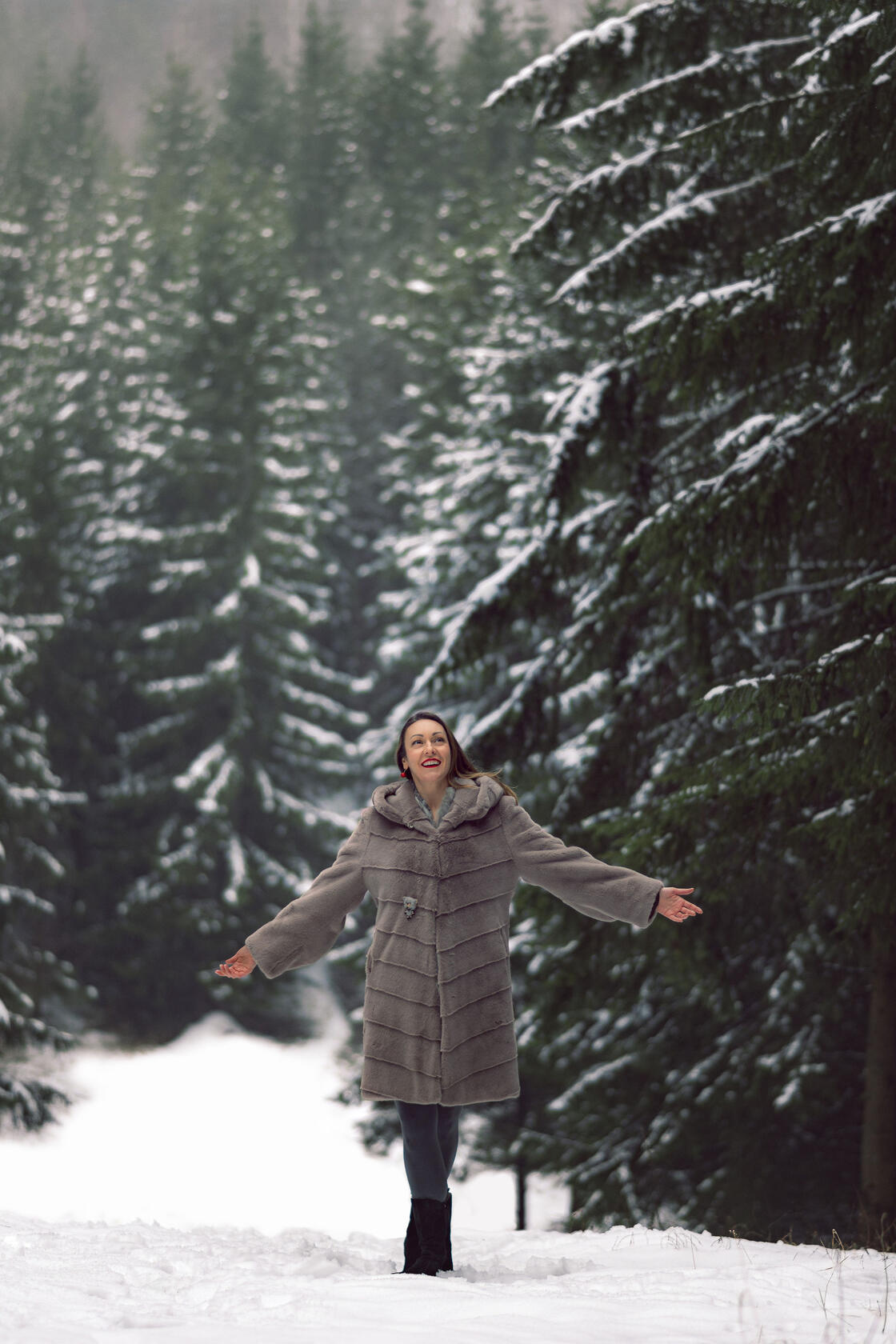 Moteris raudonomis lūpomis apsivilkusi kailinius gėrisi gamta, už jos apsnigtas spygliuočių miškas