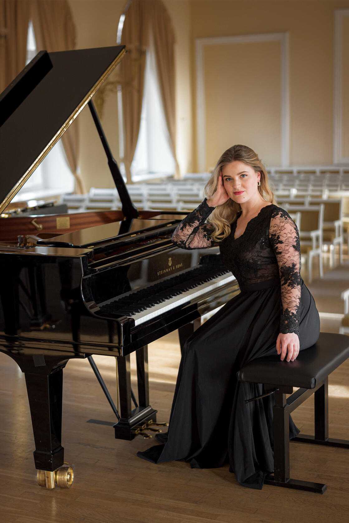 Muzikantė su suknele sėdi prie fortepijono, žvelgia į objektyvą. Vidutinio plano kadras