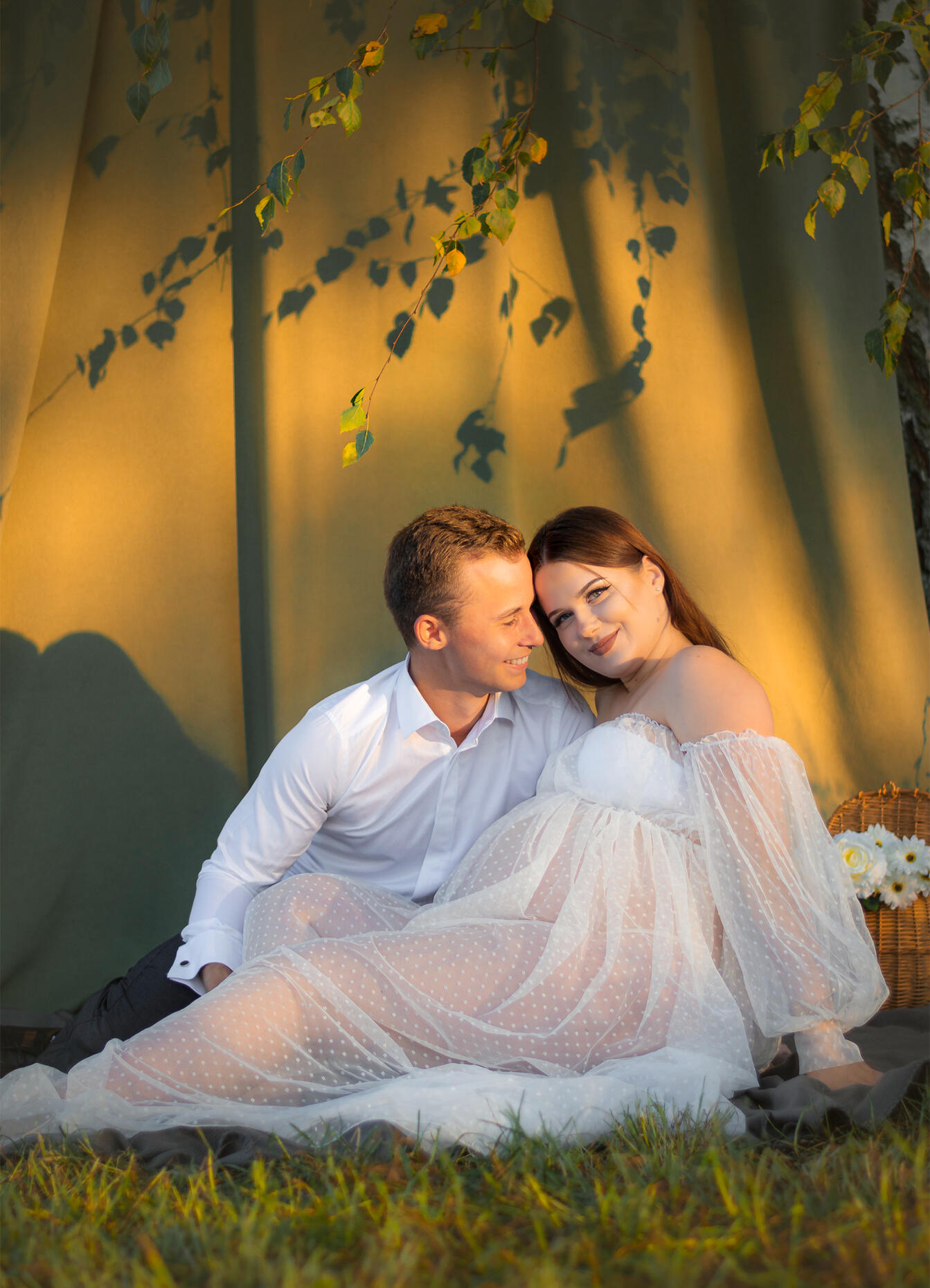 nėščia moteris su partneriu laimingi sėdi tarp žaliuojančių beržo šakų, į juos šviečia vakarėjanti saulė, jie pozuoja