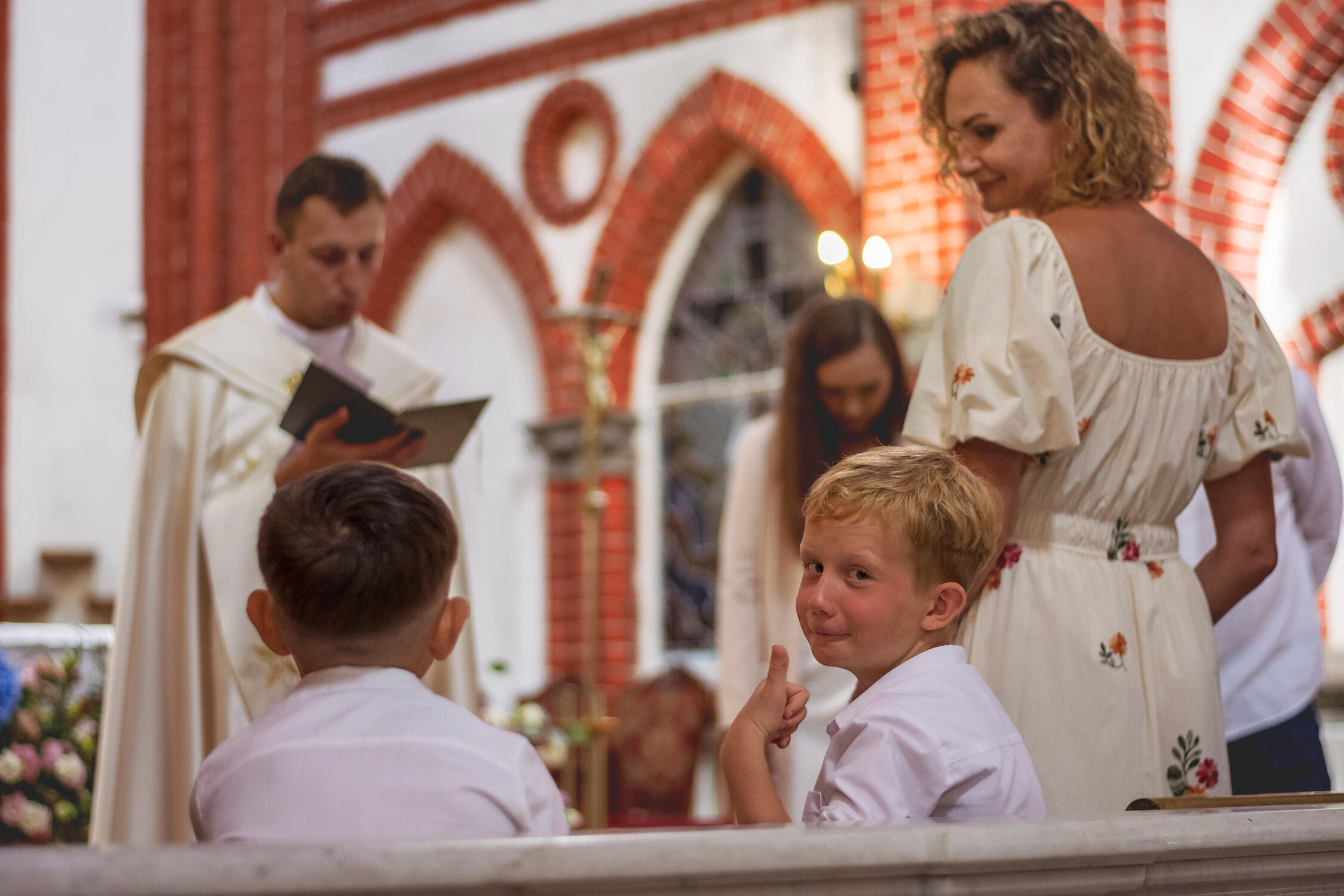 Krikštynų fotosesija. Krikštynos liuks! Vyksta ceremonija, berniukas atsisukęs į fotografą rodo liuks